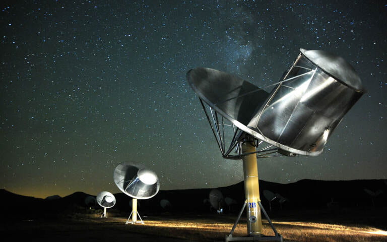 photo showing the allen telescope array. three radio dishes are shown in a line lit up on a dark night, with many stars visible in the sky